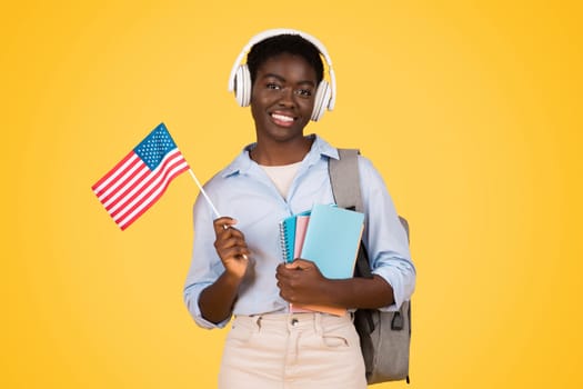 Happy young african american woman student in wireless headphones, with books, flag of USA