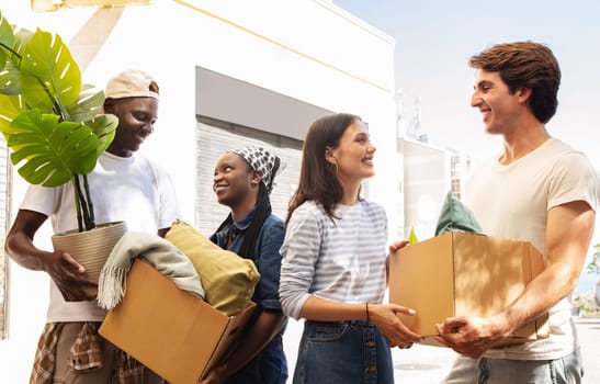 Relocation, real estate concept. Positive cheerful young multiracial people friends two ladies and two guys moving to new house, holding cardboxes with belongings stuff, plants, standing on street