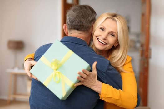 Emotional mature woman hugging husband holding gift, celebrating anniversary indoor