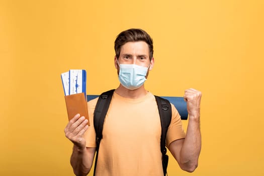 Male tourist with backpack wearing medical mask, holding tickets with passport and clenching fist on yellow background