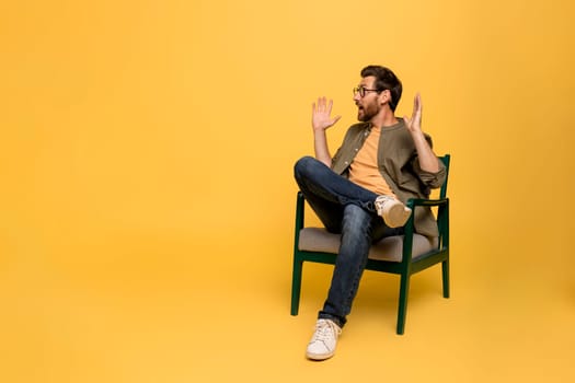 Shocked middle aged man opening mouth in amazement looking aside at free space sitting in chair over yellow background