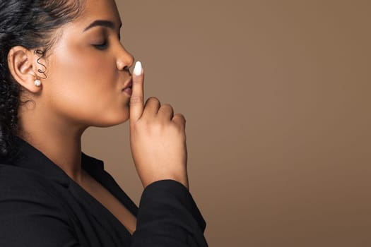 Keep silence. Latin woman showing shh gesture, holding finger near lips, brown background, side view, copy space