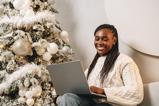 Beautiful young african female sitting with laptop near Christmas tree at home