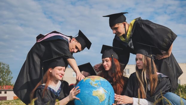 Graduates in black robes examine a geographical globe sitting on the grass.