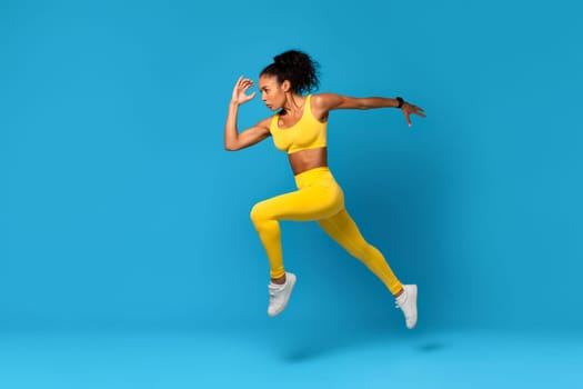 African Lady In Fitwear Exercising Jumping Over Blue Studio Background