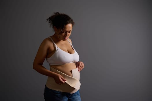 Beautiful confident woman, young mother putting on elastic bandage after c-section, isolated on gray studio background