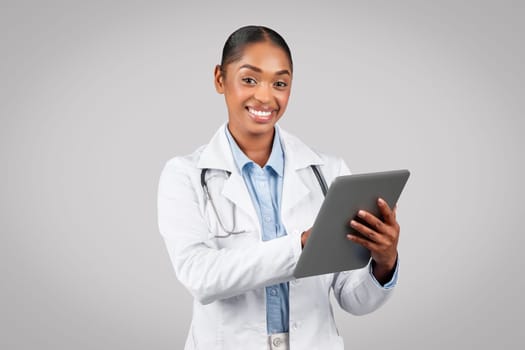 Positive young black woman doctor in white coat, chatting at tablet, typing