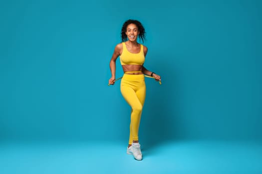 Happy black woman in sportswear measuring waist with tape, studio