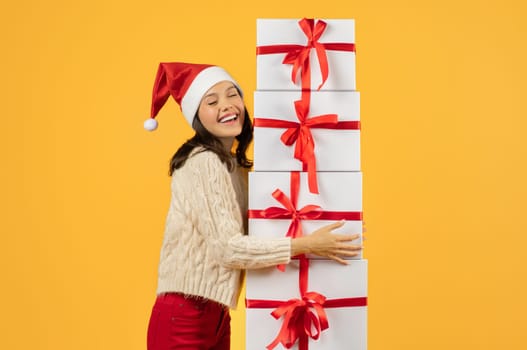Cheerful woman embracing stack of Christmas gifts on yellow backdrop