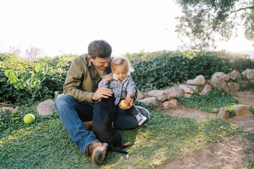 Dad with a little girl on his knees sits on the grass and pets a puppy. High quality photo