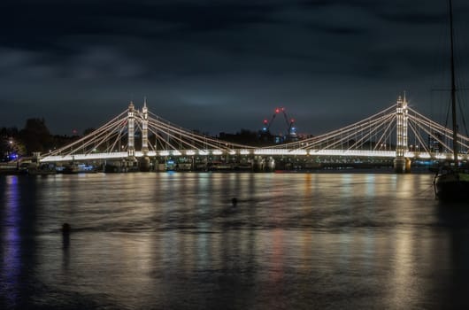 Nocturnal view of floodlit Albert Bridge from Chelsea Bridge with the reflection of the light in very calm water.