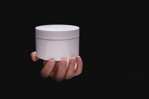White cosmetic packaging mockup. A woman's hand holds a round jar of body cream on a black background. Beauty industry concept. Close-up, copy space, banner