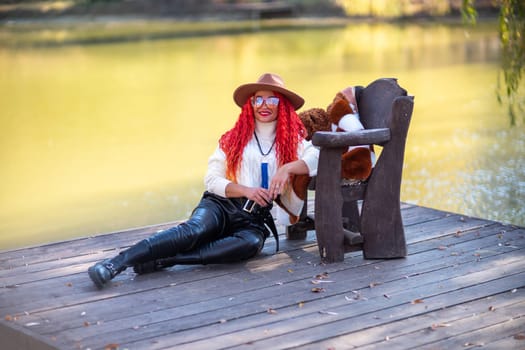 Autumn lake woman. In autumn, she sits by the pond on a wooden pier and admires nature with red hair and a hat. Tourism concept, weekend outside the city.