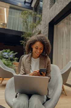 Positive female freelancer holding mobile phone while sitting in coworking and work on laptop