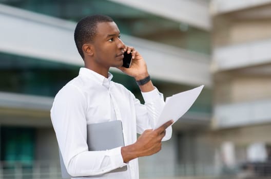 Serious black businessman holding papers and talking on cellphone while standing outdoors