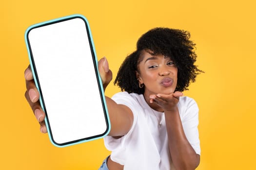 Black woman holding huge smartphone playfully blowing a kiss, studio