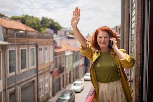 Cheerful senior woman standing on balcony, waving hand and talking on cellphone