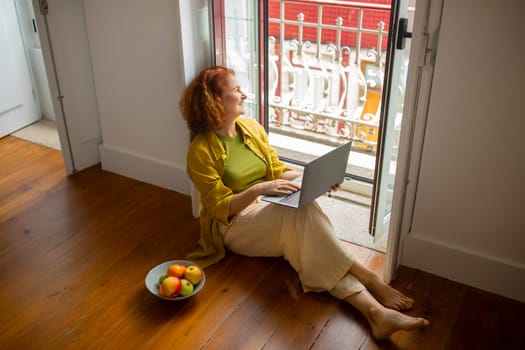 Senior Woman Relaxing With Laptop On Floor Near Open Window At Home
