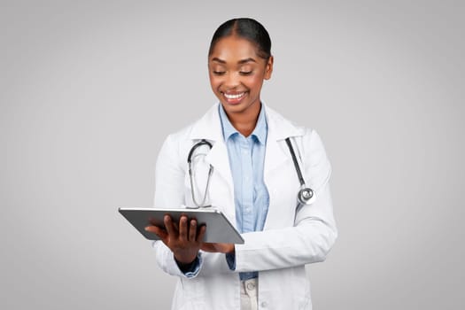 Busy smiling young black woman doctor in white coat typing at tablet, work at device