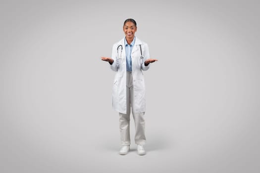 Smiling black young lady doctor in white coat, hold empty space on hands