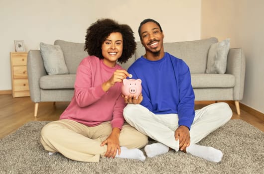 Cheerful black spouses holding and putting money in piggybank indoor