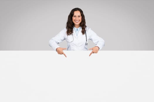 Free checkup. Happy doctor woman in medical uniform showing white blank placard for advertisement or text, mockup