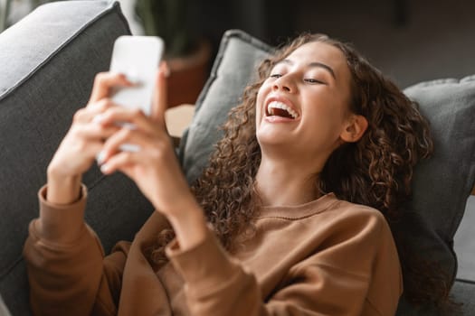 Joyful teenage girl texting on smartphone lounging on couch indoors
