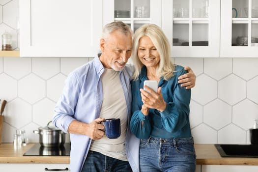 Smiling Elderly Husband And Wife Relaxing In Kitchen With Smartphone And Coffee