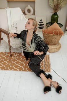 Woman glamorous pajamas. A beautiful blonde in black pajamas is sitting relaxed in a chair in black pajamas with feathers. Beige stones are all over the interior.