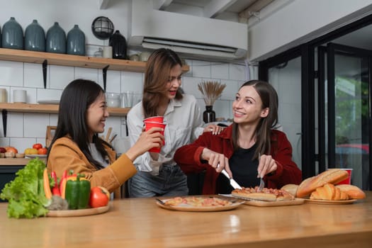 A group of young female friends have a party with pizza on the table and red drink glasses. Talk and live together happy, having fun at home.