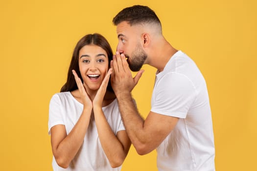 Happy young european man in white t-shirt whispers in ear of surprised lady secret