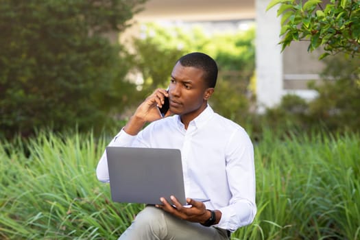 Multitasking Concept. Black Young Businessman Talking On Cellphone And Using Laptop Outdoors