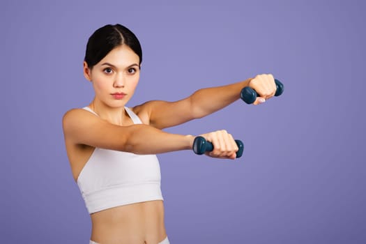 Serious caucasian slim woman in sportswear doing exercises with hand dumbbells, looking at camera, purple background
