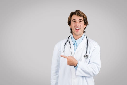 Joyful man doctor pointing to side, stethoscope around neck on grey backdrop