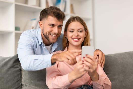 Young spouses using mobile phone together, scrolling through apps and websites, pointing and sharing online finds, sitting together on couch at home, engaged with digital device
