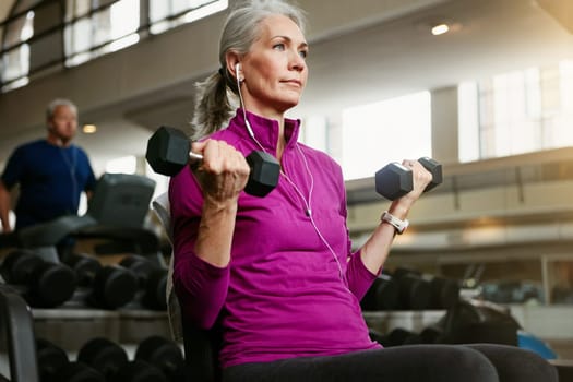 Old woman, fitness and dumbbells at gym for training, wellness and cardio with earphones, music or mindset. Weightlifting, bodybuilding and senior female person at a sports center for biceps workout