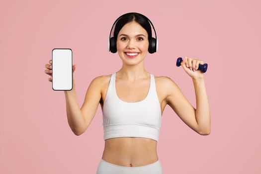 Cheerful european lady in sportswear, headphones with dumbbell showing cellphone blank screen, mockup