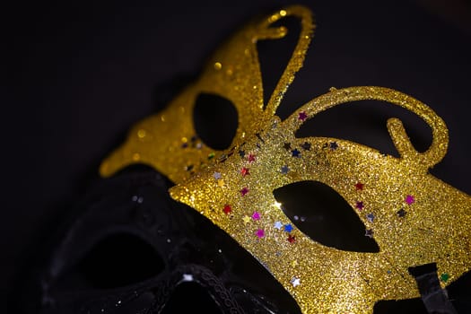 A close-up photo of two carnival masks, one gold and one black, on a black background. The gold mask is decorated with colorful rhinestones.