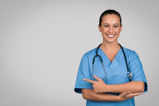 Positive nurse lady in blue workwear advertising space for advertisement, pointing finger aside, medical offer text