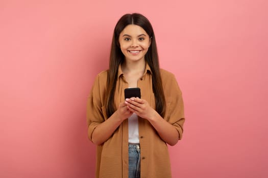 Portrait Of Smiling Teen Girl With Smartphone In Hands Standing Over Pink Background In Studio, Happy Cute Female Teenager Browsing New App On Mobile Phone Or Messaging With Friends, Copy Space