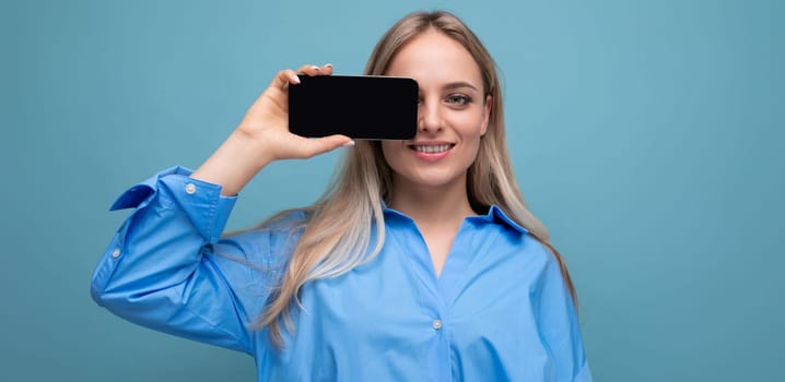 attractive confident blond girl holding a smartphone with a screen at the camera with a blank mockup for a website on a blue background