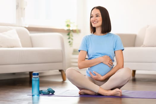 Expectant woman exercising sitting in lotus pose on floor indoors