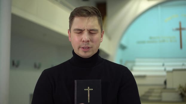 A young man prays holding a bible in his hands in a church against the background of a cross. A Protestant man stands in a church and prays.