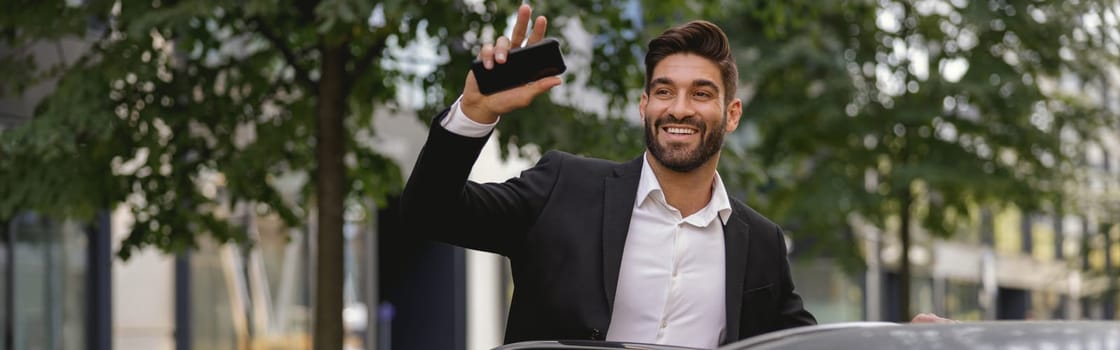 Cheerful businessman standing near car with mobile phone and waving hi to colleagues