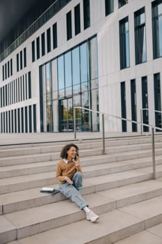 Cheerful business woman talking phone with client while sitting on stairs on building background