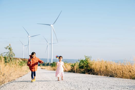 Two children girl with pinwheel playfully run by windmills