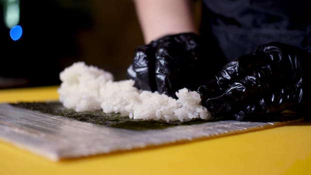 The chef putting rice on the nori sheet, Japanese cuisine concept. Close up for hands in black cooking gloves touching white prepared rice for making rolls.