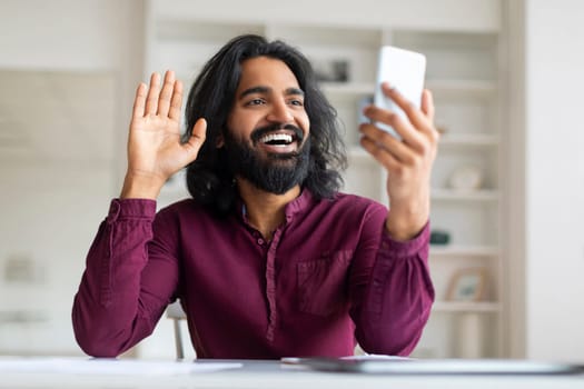 Smiling Indian freelancer man waving hand while having video call on smartphone