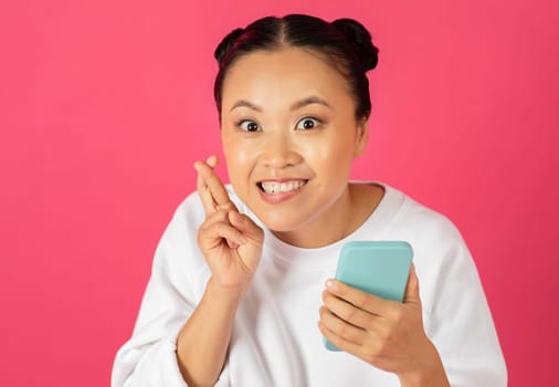 Cheerful Asian woman crossing her fingers for good luck and holding smartphone