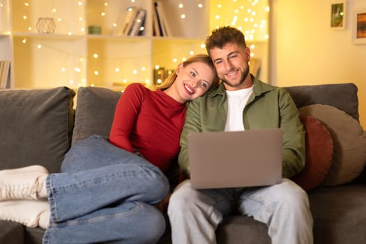 Couple with laptop enjoying time on couch, festive lights backdrop